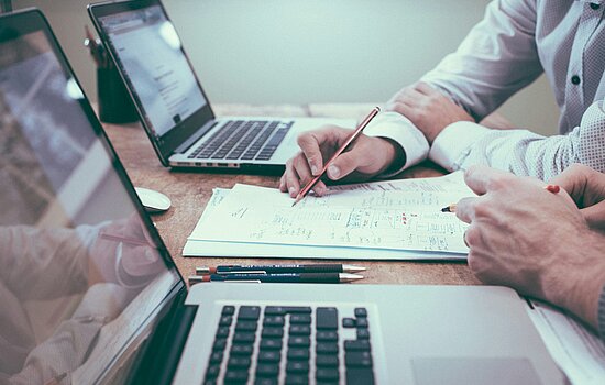 Zwei Personen arbeiten an Laptops, überprüfen Papiere und machen Notizen. Fokus auf die Hände, die einen Bleistift und einen Stift halten.