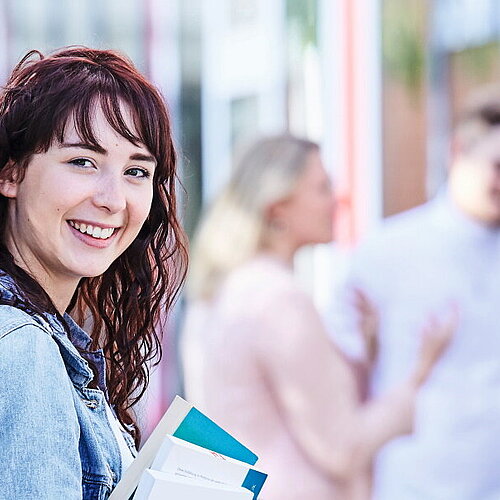 Public Management studieren Junge Studentin mit lockigem Haar, Jeansjacke und Rucksack hält Bücher in der Hand und lächelt, während im Hintergrund Studierende miteinander sprechen.
