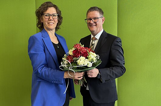Amtsantritt Kanzler Markus Dammler Rektorin HVF Rauskala und Kanzler Dammler mit Blumenstrauß vor grüner Wand.