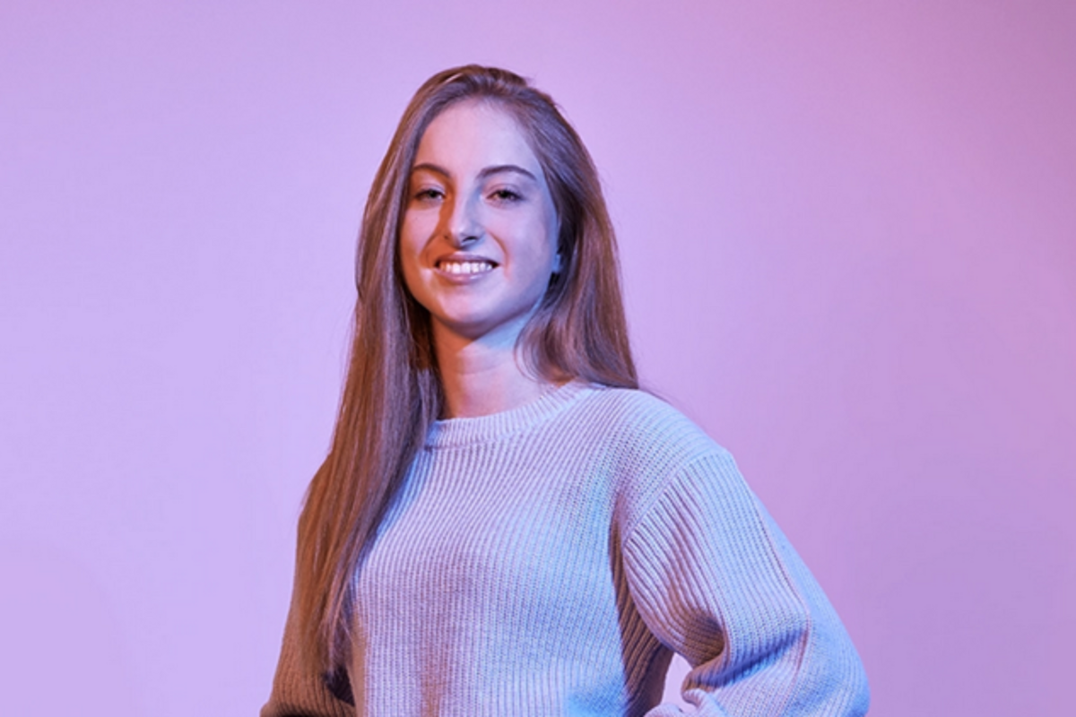 Rentenversicherung studieren Porträt einer jungen Studentin der Rentenversicherung mit langen Haaren und Strickpullover vor einem lila Hintergrund, lächelnd und mit selbstbewusster Haltung.