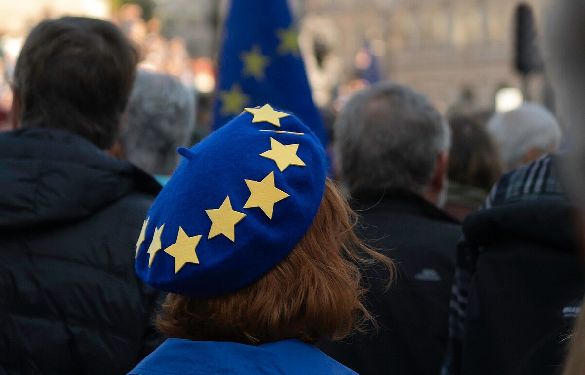 Master Europäisches Verwaltungsmanagement studieren Frau mit blauem EU-Barett in einer Menschenmenge bei einer pro-europäischen Demonstration auf dem Trafalgar Square in London, im Hintergrund sind Gebäude und eine EU-Flagge sichtbar.