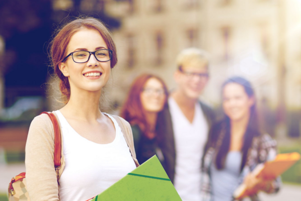 Master Public Management studieren Junge Studentin mit Brille und Ordnern lächelt in die Kamera, im Hintergrund weitere Studierende auf einem sonnigen Campus.