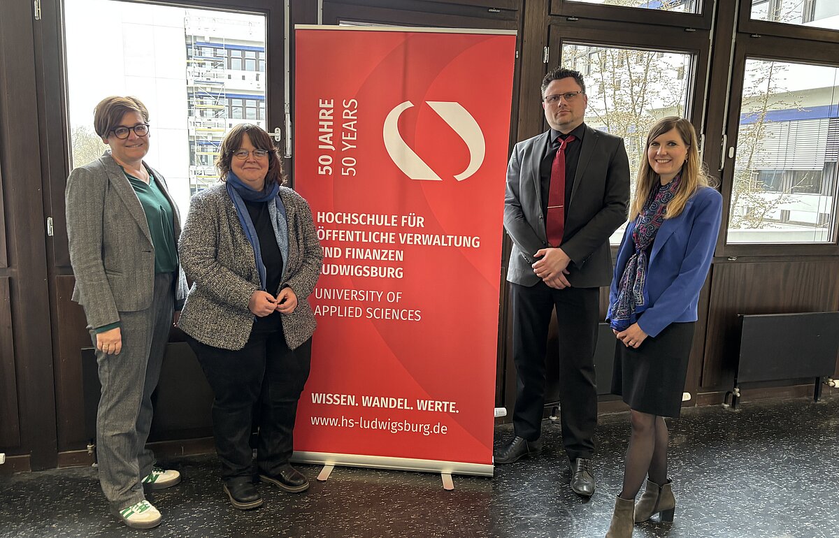 Gruppenfoto mit Abgeordneten an der Hochschule für öffentliche Verwaltung und Finanzen Ludwigsburg Vier Personen stehen vor einem roten Roll-up der Hochschule für öffentliche Verwaltung und Finanzen Ludwigsburg mit der Aufschrift "50 Jahre – 50 Years". Die Personen sind geschäftlich gekleidet und lächeln in die Kamera. Im Hintergrund sind große Fenster mit Blick auf ein modernes Gebäude zu sehen.